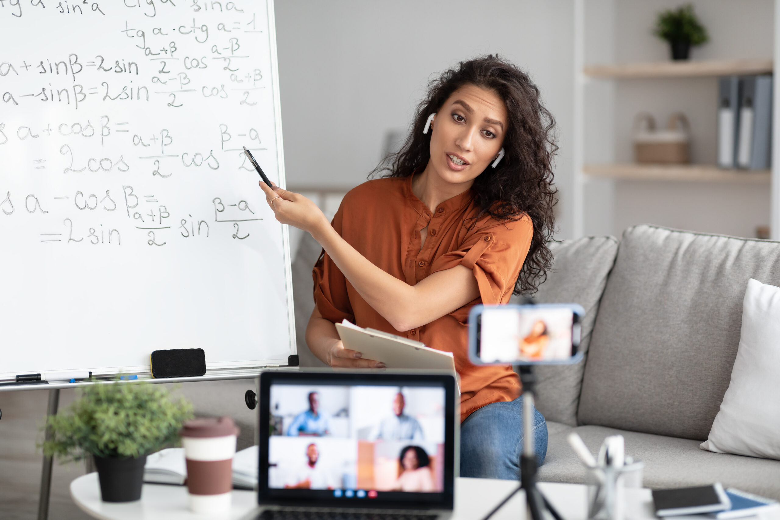 Online Lesson Concept. Portrait of young tutor pointing at board with trigonometric functions, explaining math to students making videochat. Woman in wireless headphones filming class on smartphone